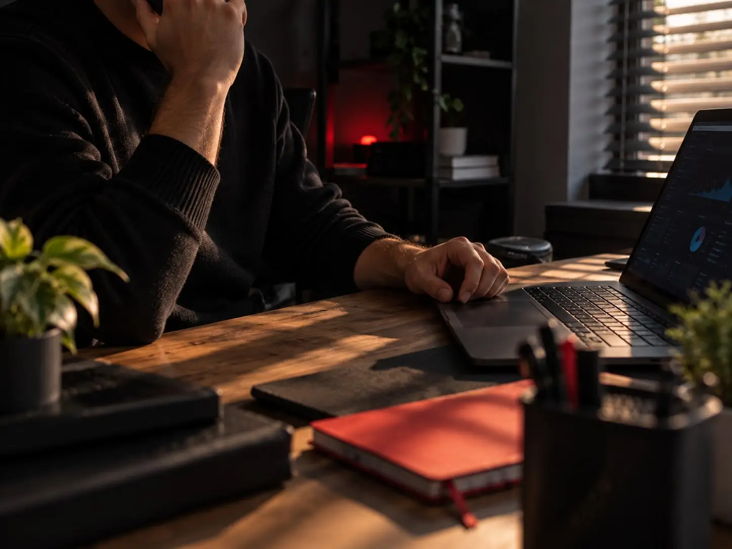 Personne en pull noir assise à son bureau, main près du visage, ordinateur portable affichant un tableau de bord et carnet rouge au premier plan.