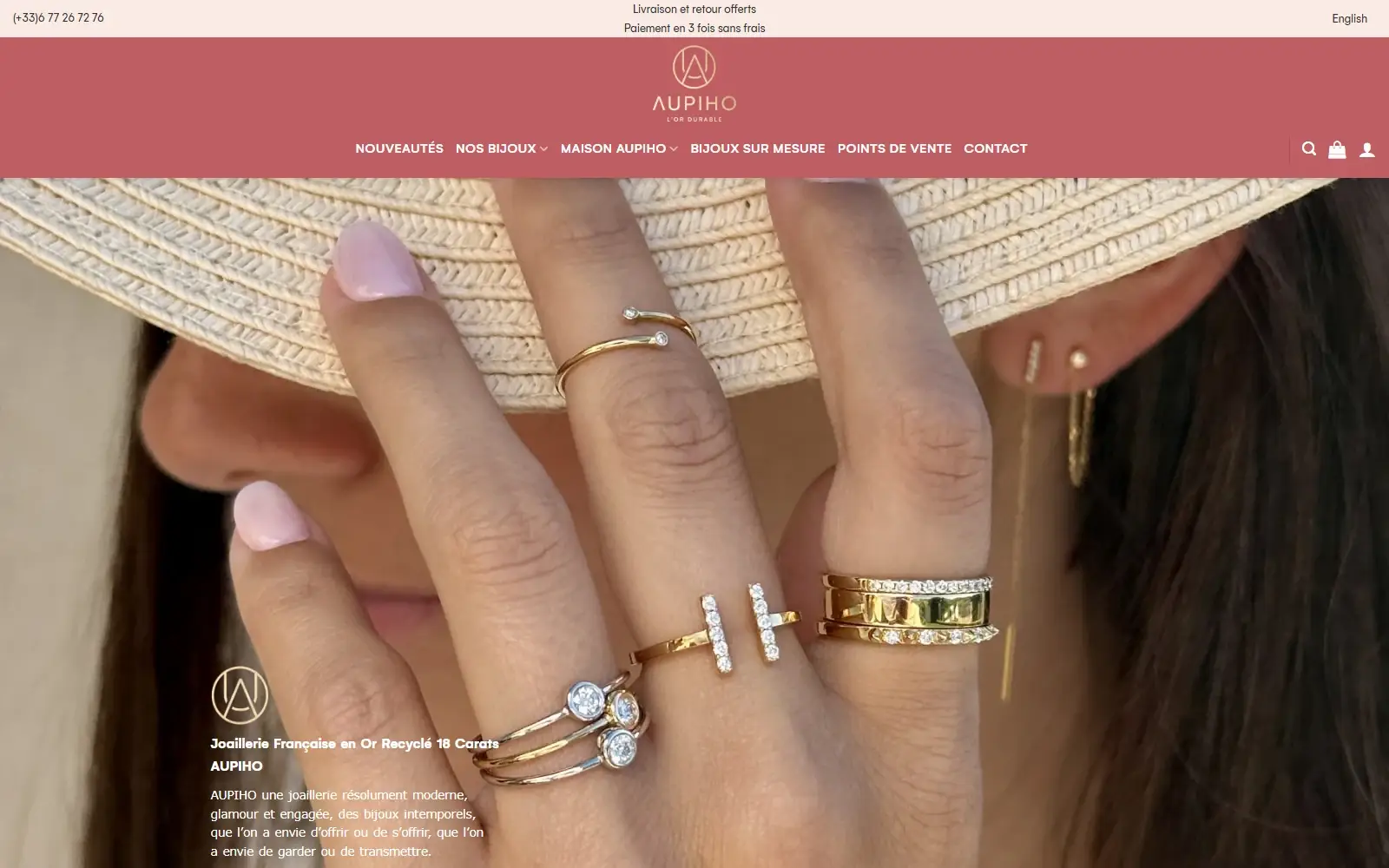 Page d’accueil d’aupiho.com : bandeau rose avec le logo AUPIHO et le menu Nouveautés, Nos bijoux, Bijoux sur mesure ; photo d’une main portant des bagues en or sous un chapeau de paille.