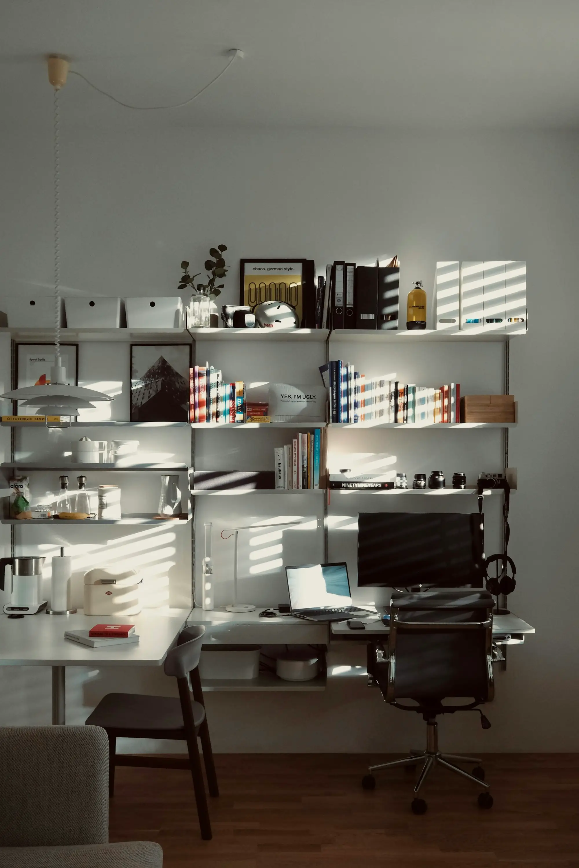 Bureau à la maison : ordinateur portable et écran sur une étagère murale, lumière du soleil à travers les stores.